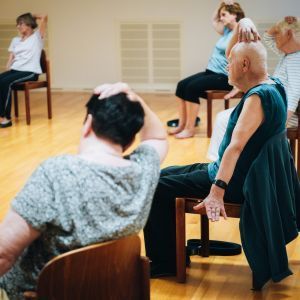 M&auml;nner und Frauen sitzen im Kreis auf einem Stuhl und dehnen ihren Nacken